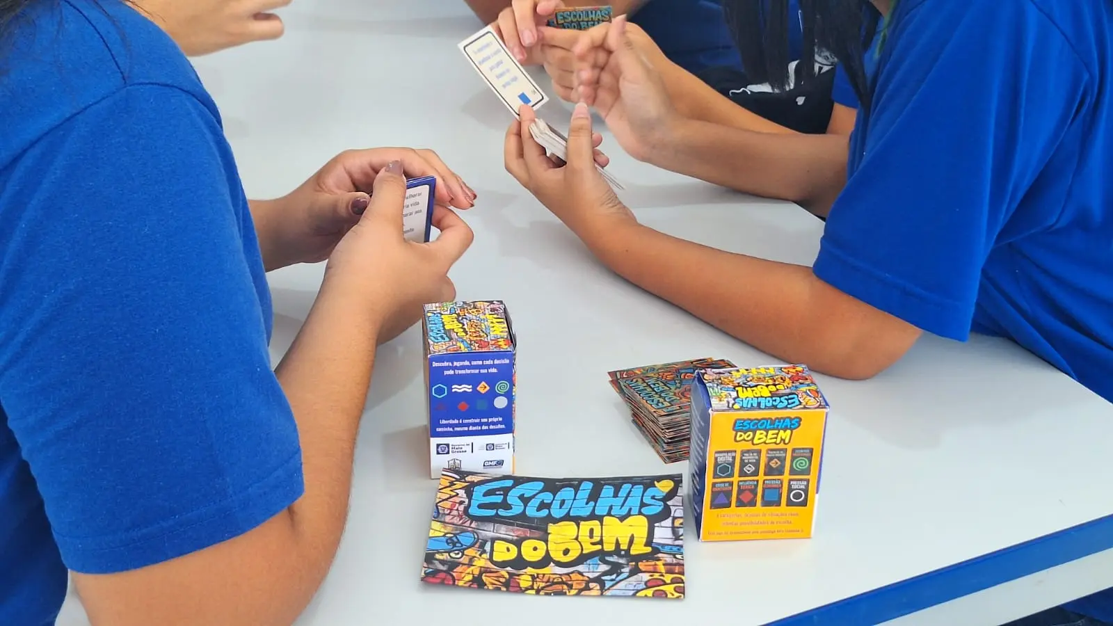 Foto colorida que mostra detalhe das mãos de alunos da rede estadual jogando o jogo de cartas Escolhas do Bem.