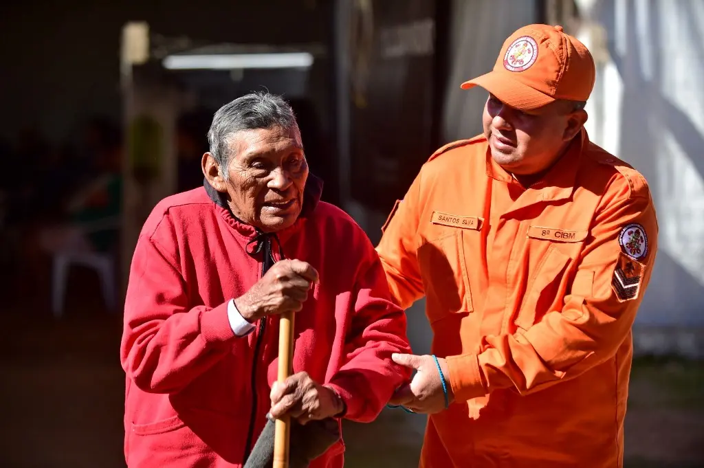 Bombeiro militar, usando uniforme laranjado, ajuda idoso a caminhar. O idoso tem traços indígenas, usa um moletom vermelho e um cajado.