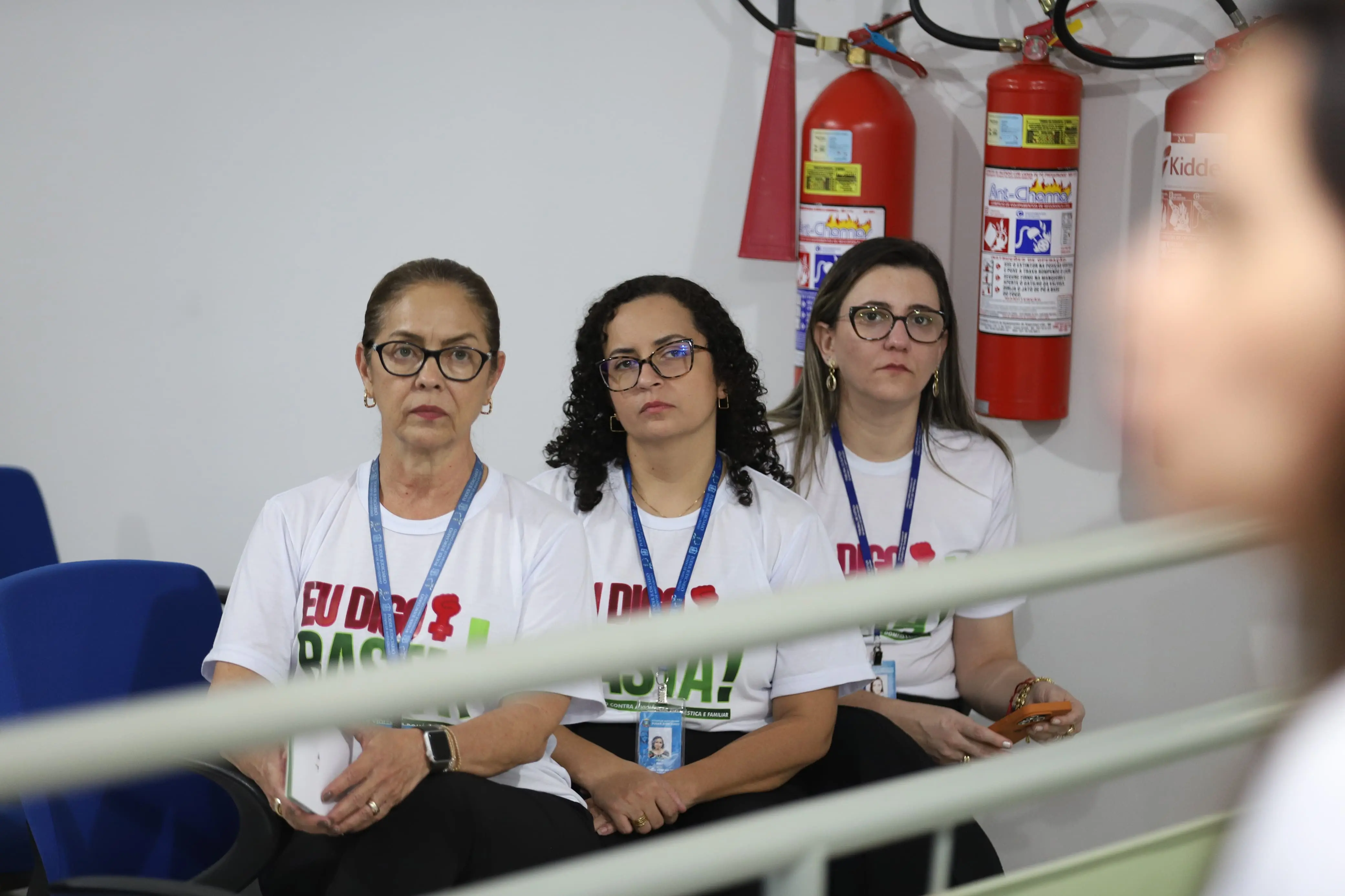 Três mulheres sentadas, com expressão séria, vestem camisetas da campanha “Eu Digo Basta!”. Usam crachás e assistem atentamente a uma atividade. Ao fundo, há extintores de incêndio fixados na parede branca.