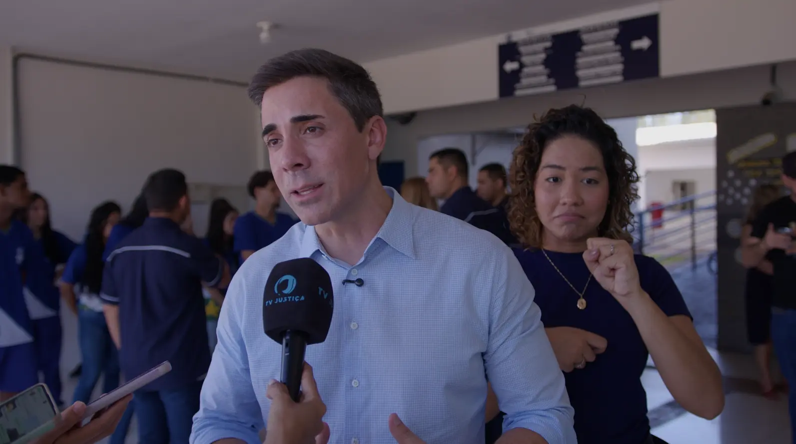 Secretário estadual de Educação, Alan Porto, durante entrevista à TV Justiça. ele é um homem branco, de cabelo castanho claro, usando camisa azul clara. Atrás dele, há uma intérprete de Libras e alunos no pátio da escola. 