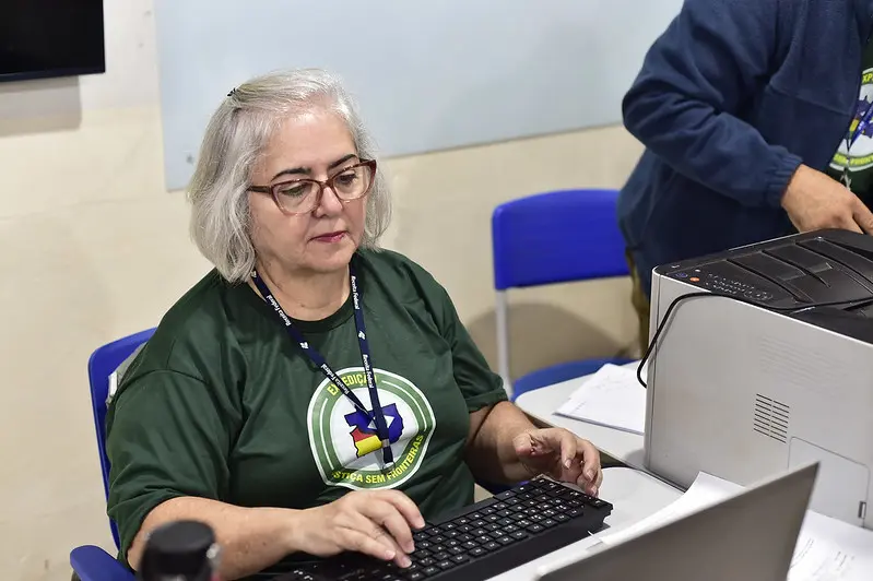 Servidora da Receita Federal trabalha em computador. Ela é uma senhora branca, de cabelos brancos, lisos e na altura dos ombros, usando óculos de grau e camiseta verde da Justiça Sem Fronteiras.