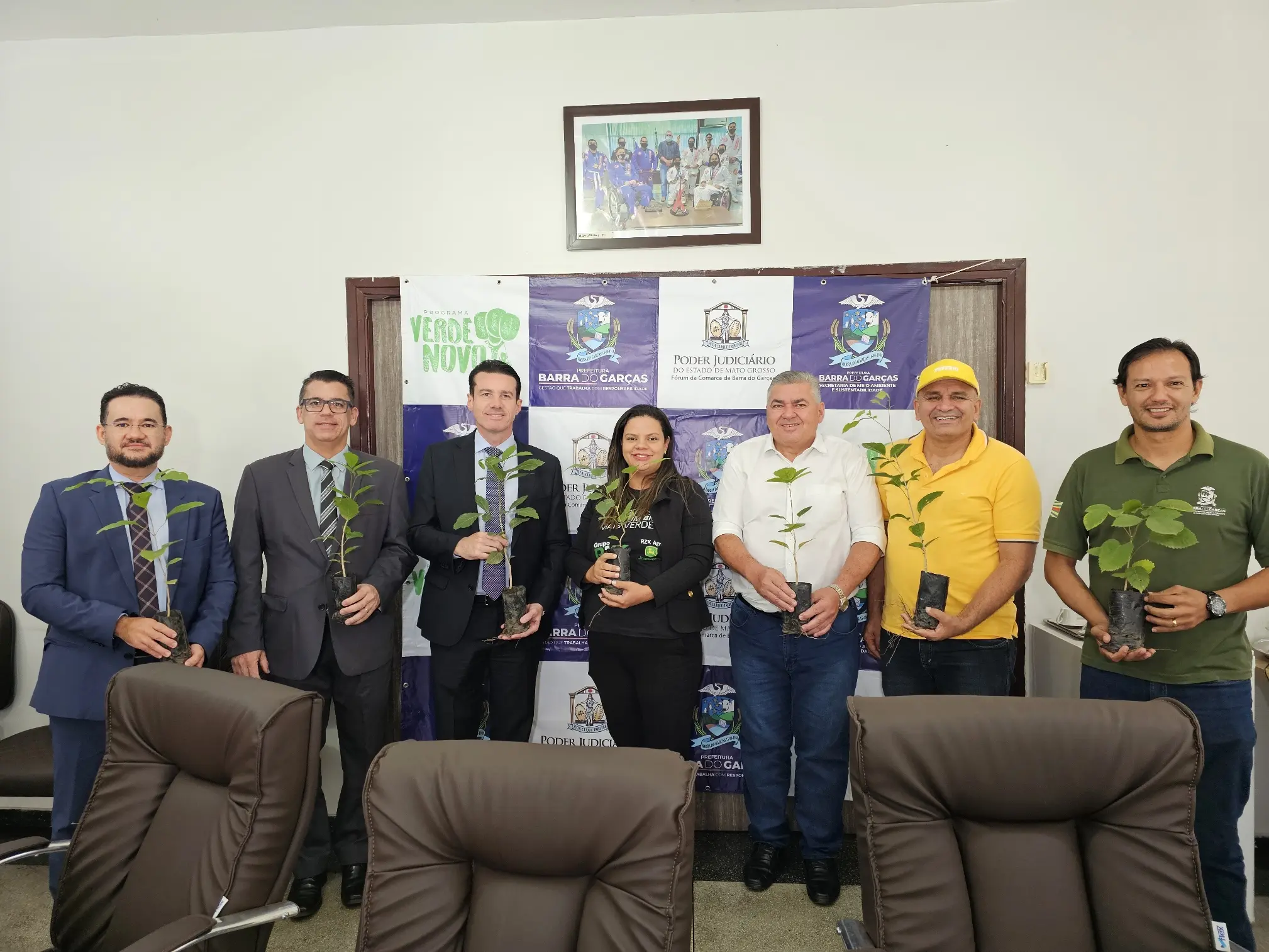 Juízes, desembargador Rodrigo Curvo, prefeito, vice-prefeito e secretário de Barra do Garças e representante da RZK Agro posam para a foto sorrindo e segurando uma muda de árvores cada um. Atrás deles, há um painel com as logos do Poder Judiciário, do Verde Novo e o brasão de Barra do Garças.