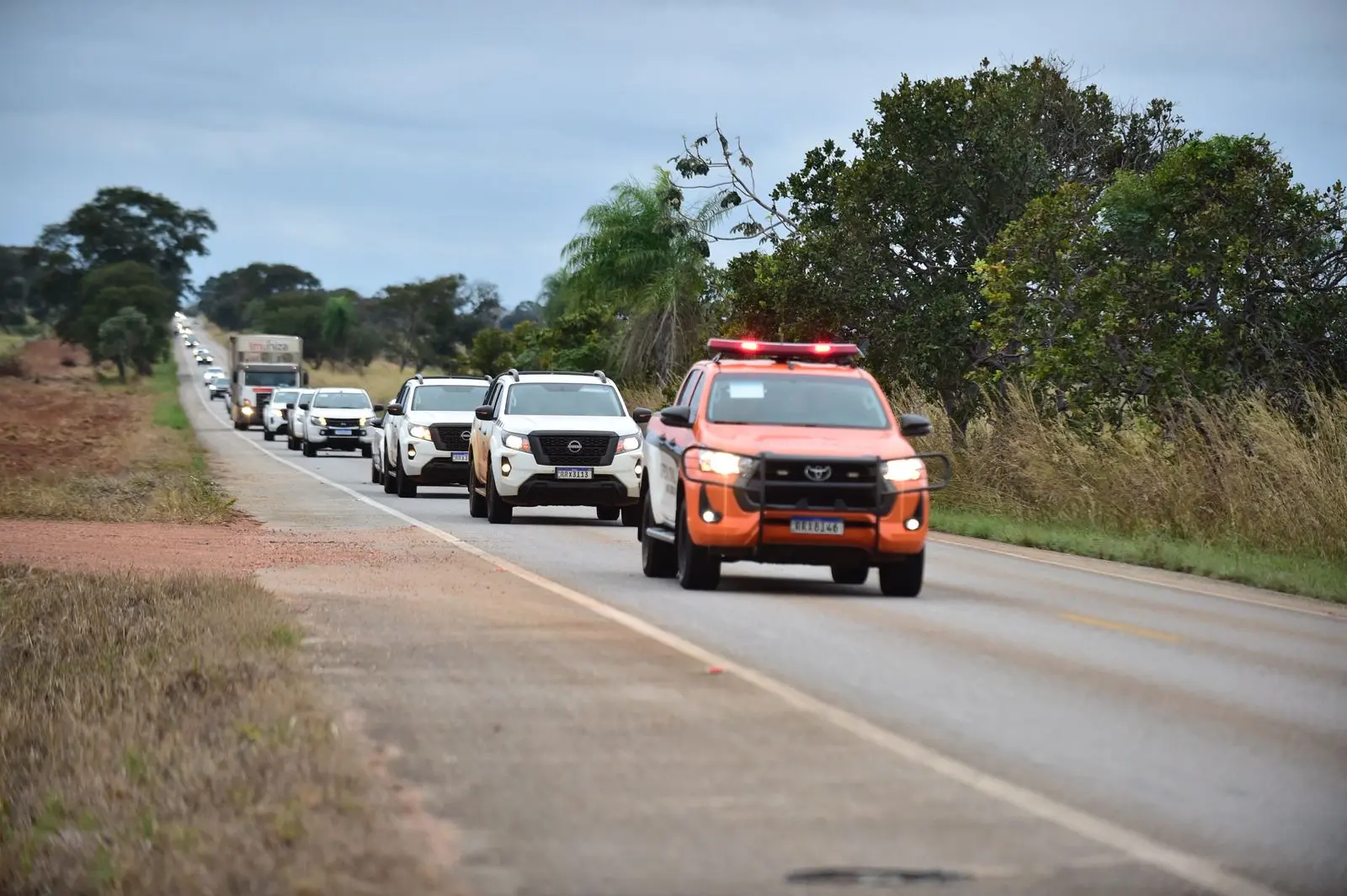 Comboio com veículos oficiais percorre estrada rodeada por vegetação e árvores. Na frente, caminhonete laranja com luzes de emergência acesas. Céu nublado e pista longa à frente.