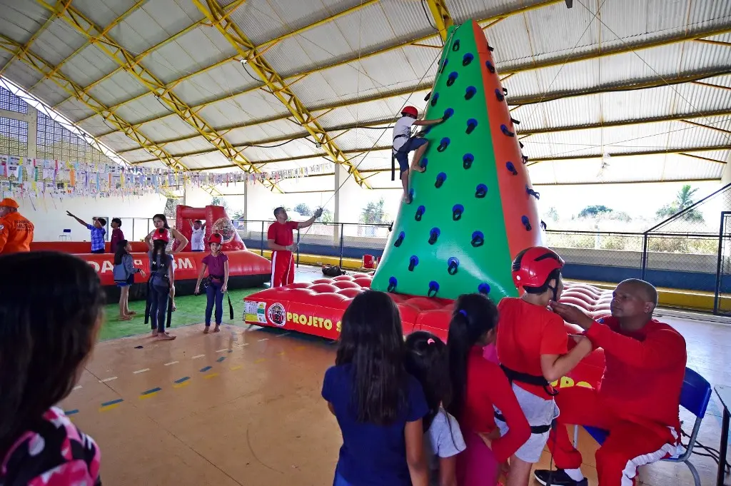 Em uma quadra esportiva, crianças brincam no campo de futebol inflável e na torre de escalada inflável do Corpo de Bombeiros. Bombeiros militares conduzem as atividades.