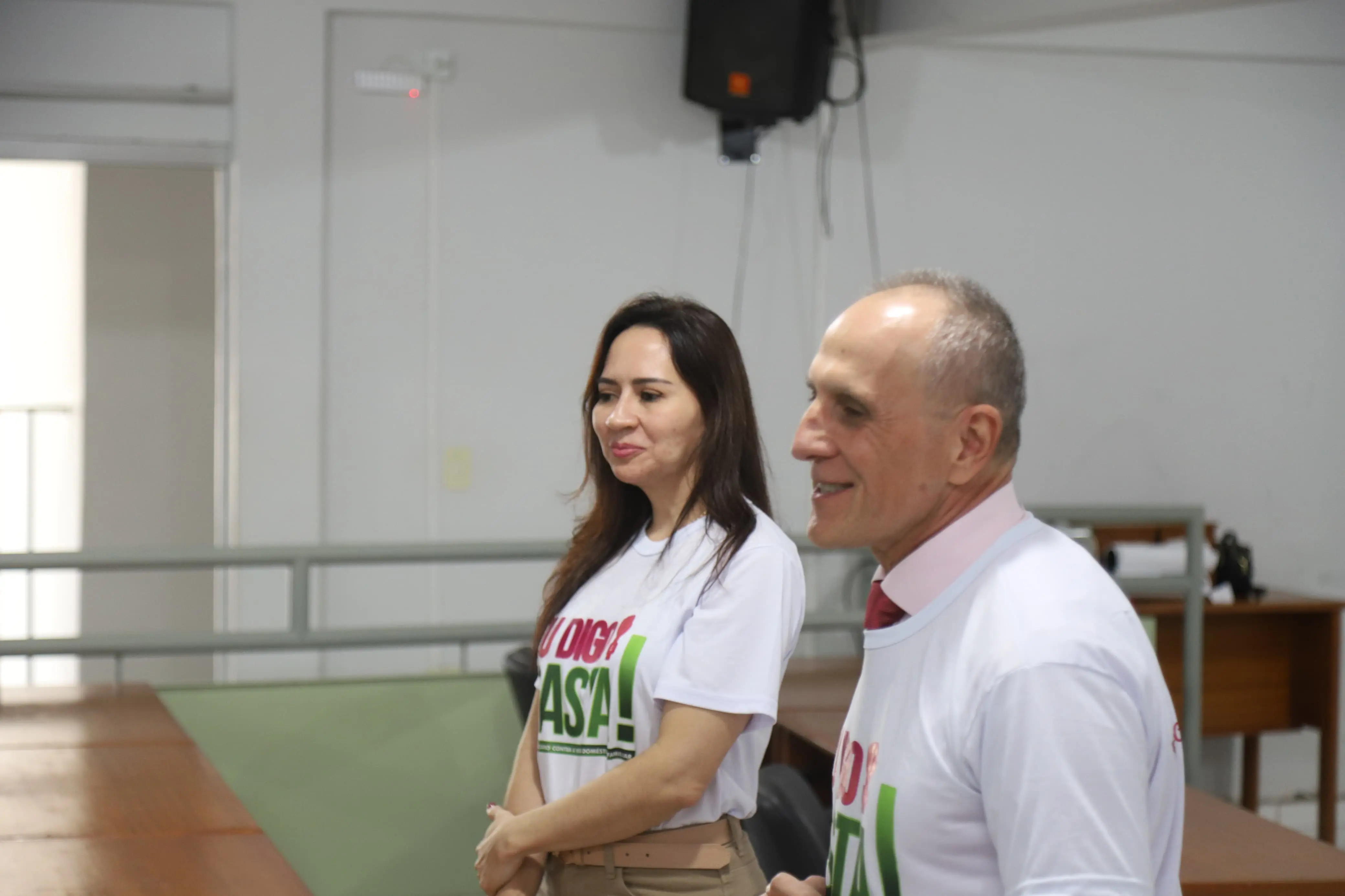 Mulher e homem sorriem e conversam em ambiente de evento. Ambos usam camisetas da campanha “Eu Digo Basta!”. Ao fundo, há mesas de madeira, paredes brancas e equipamentos de som. A imagem transmite leveza e interação.