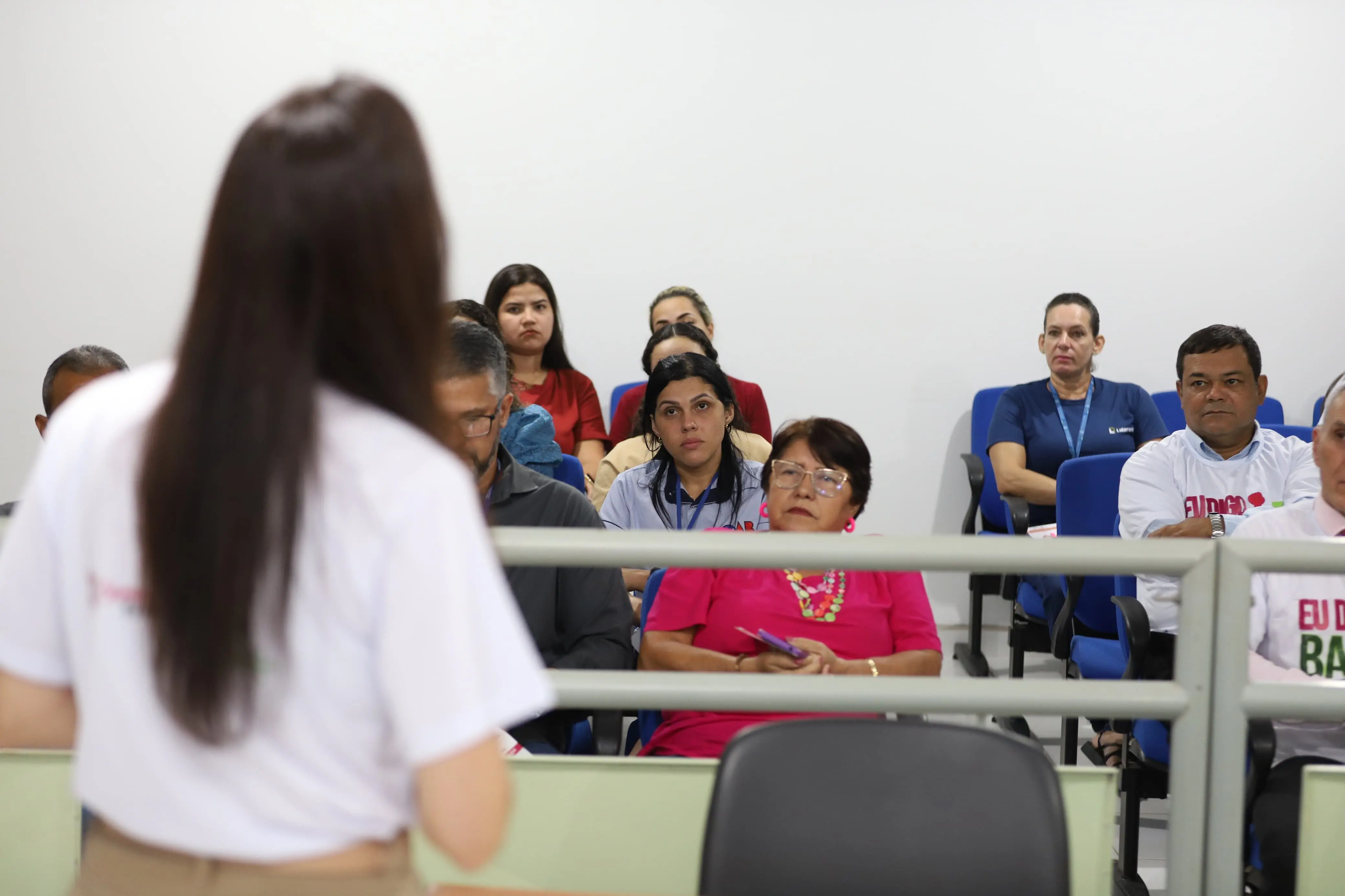 Pessoas estão sentadas em cadeiras azuis, assistindo a uma apresentação. No primeiro plano, vê-se a palestrante de costas. Alguns participantes vestem camisetas da campanha “Eu Digo Basta!”, e outros usam roupas casuais ou crachás.