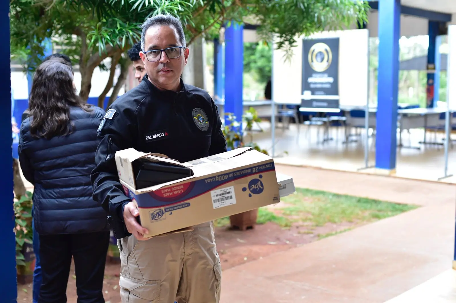 Homem uniformizado carrega caixa de papelão com equipamentos. Ao fundo, estrutura com mesas e cadeiras montada para atendimentos, sob cobertura azul e branca com árvores próximas