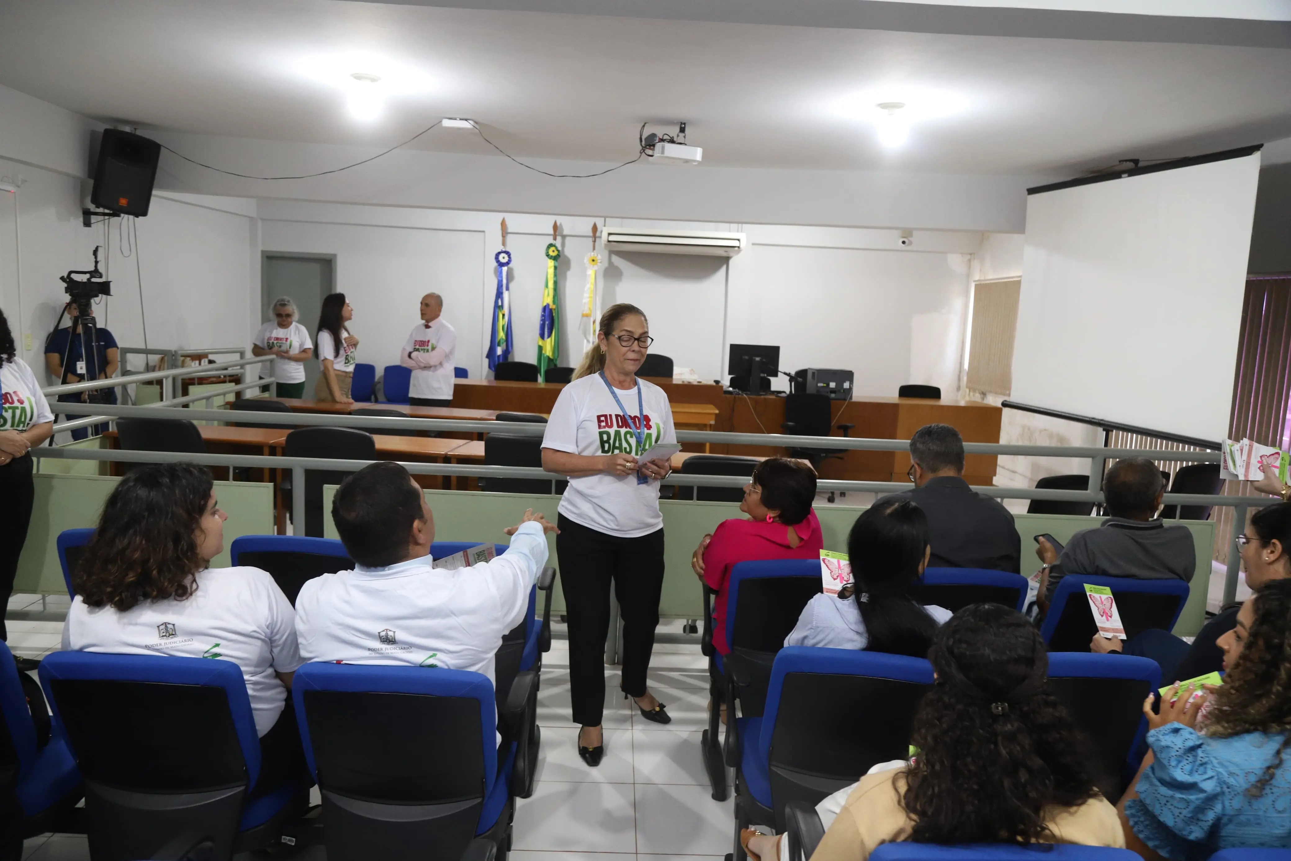Mulher distribui panfletos da campanha “Eu Digo Basta!” em plenário com público sentado. Participantes usam camisetas da ação e observam com atenção. Ao fundo, há bandeiras, projetor e equipe organizadora em pé, conversando próximo ao púlpito.