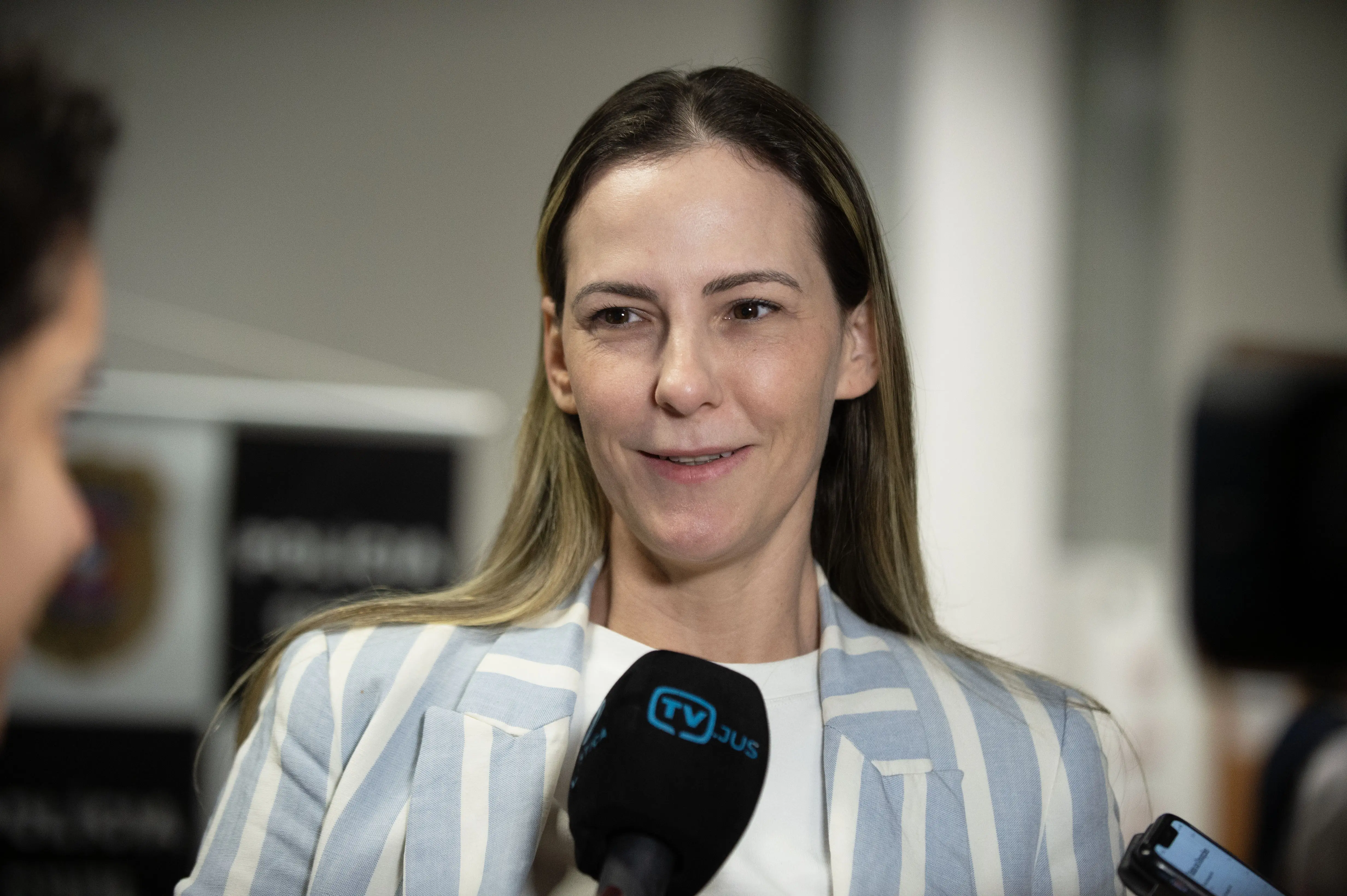Imagem horizontal colorida da advogada Daniela Felber. Ela é uma mulher branca, magra, loira, de cabelos compridos, lisos. Usa uma blusa branca e blaser listrado branco e azul claro