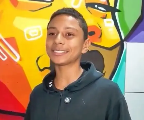 O estudante Leonardo está olhando e sorrindo para foto. Ele é magro, negro, cabelos curtos e usa um casaco preto